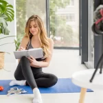 A woman with long blonde hair writes in a notebook on a yoga mat. A potted plant and camera on a tripod are nearby.