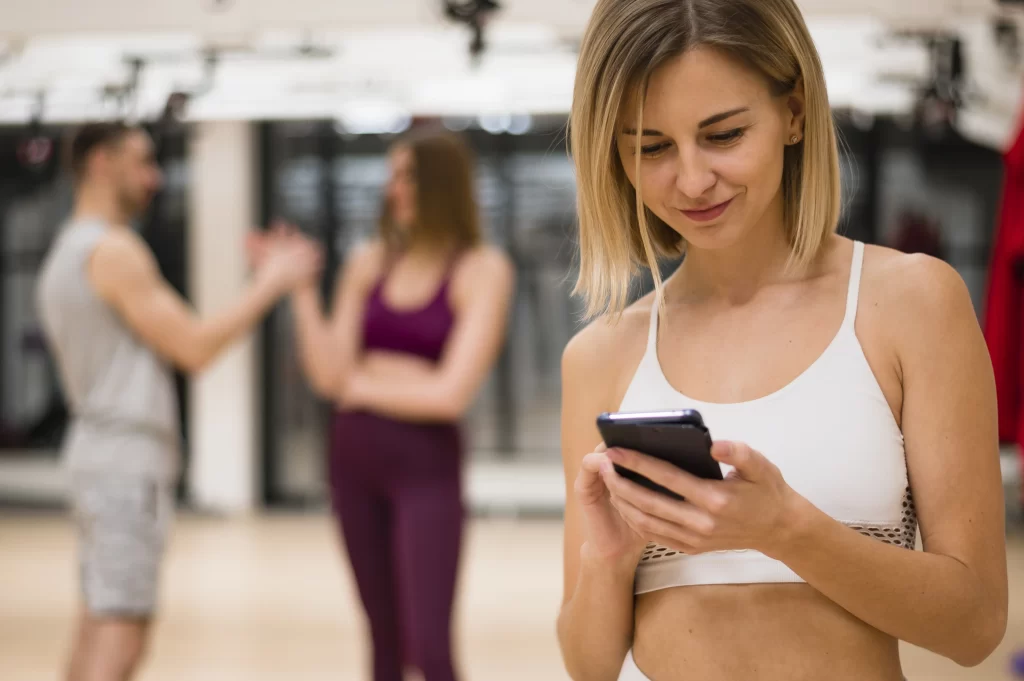A young woman in a white sports bra smiles while looking at her phone. Two blurred people are behind her.