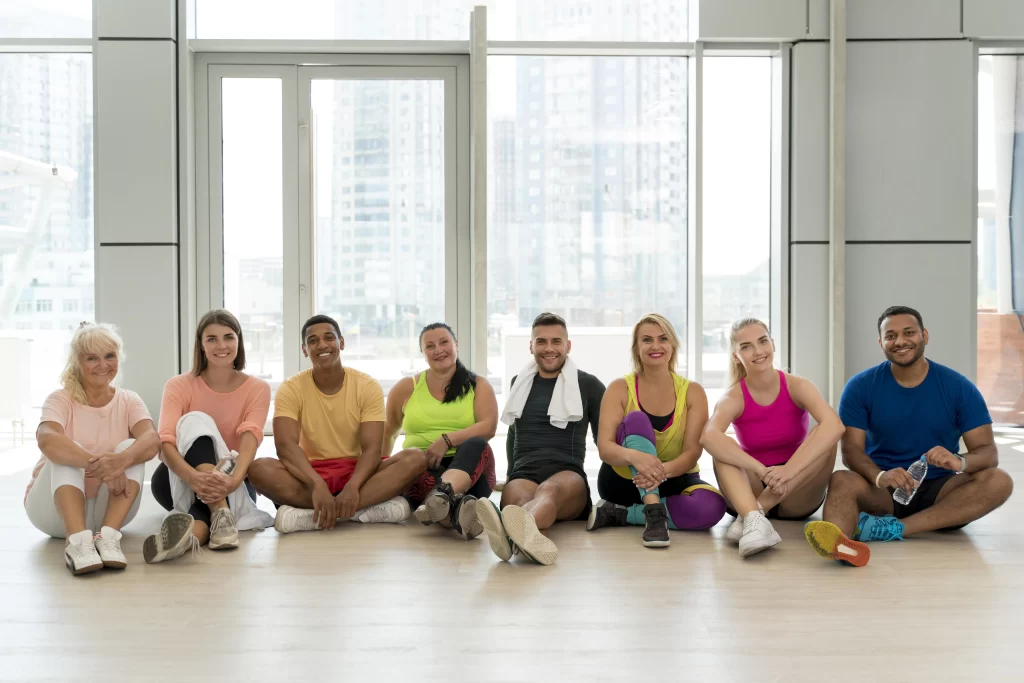 A diverse group of eight smiling people in athletic wear sits on a wooden floor in a bright studio.