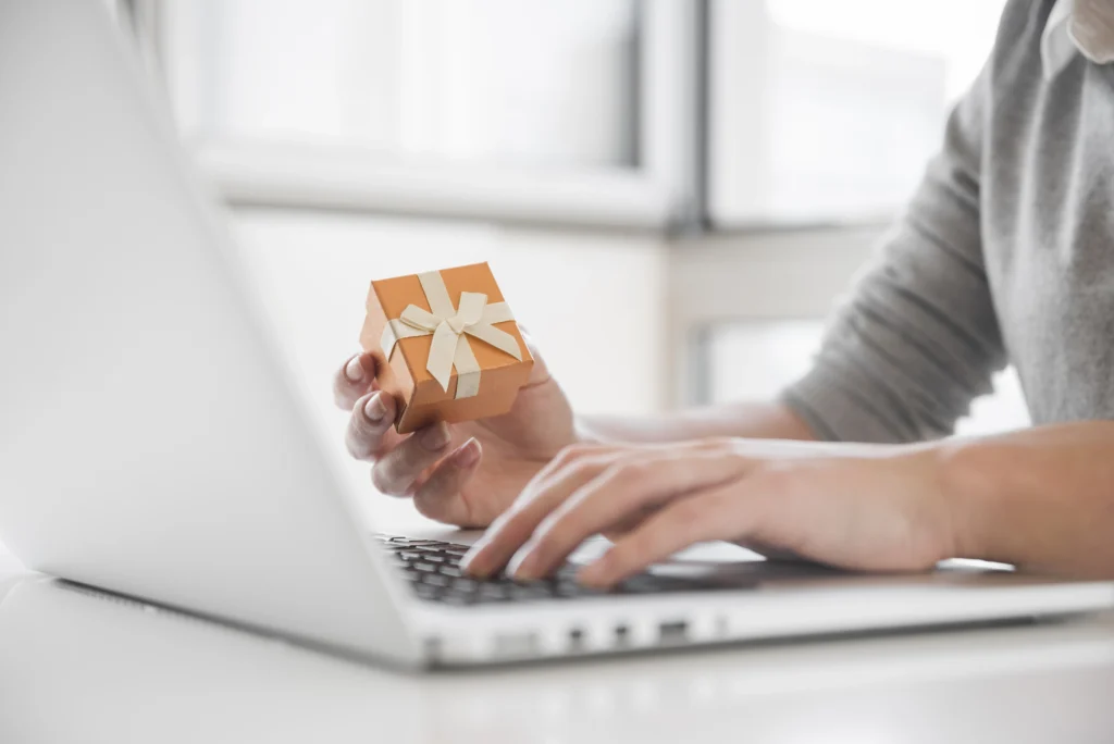 Person in gray sweater holding orange gift box with cream ribbon while typing on laptop at desk near bright window.