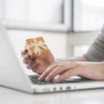 Person in gray sweater holding orange gift box with cream ribbon while typing on laptop at desk near bright window.