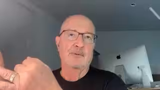 Screenshot of a bald man with a mustache and glasses wearing a black shirt, who looks up and gestures with his left hand.