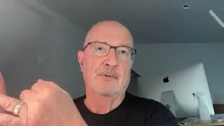 Screenshot of a bald man with a mustache and glasses wearing a black shirt, who looks up and gestures with his left hand.